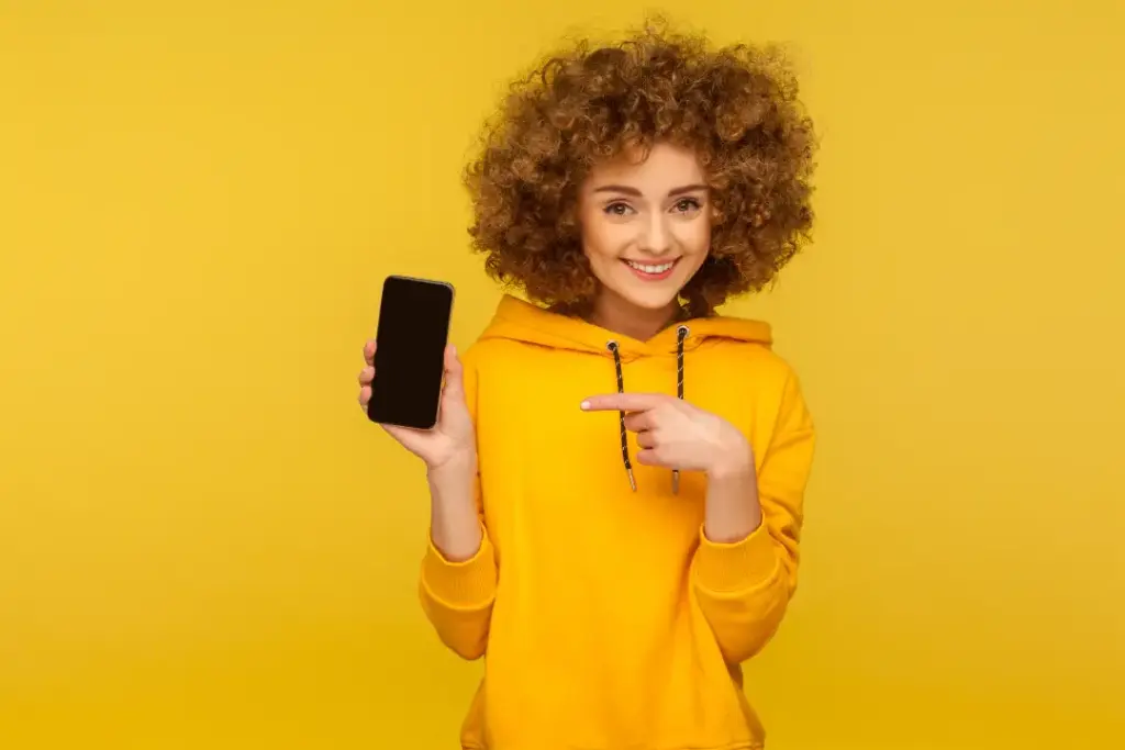 Gelber Hintergrund. Junge Frau mit gelbem Hoodie zeigt lächelnd mit dem linken Zeigefinger auf Ihr Handy in der rechten Hand.